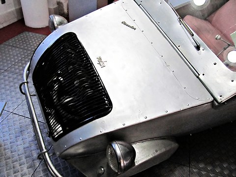 Looking down on a riveted silver aluminium bonnet with a Biscuter script and a dark curved grille, tubular bumper below. Opens a larger view