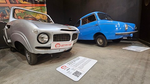 Silver open car with twin mesh grilles seen from the front left, a blue bubble-shaped microcar behind it, information board on the floor. Opens a larger view