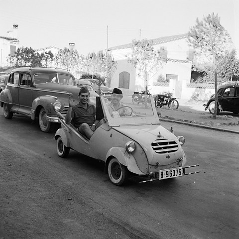 Black-and-white photograph of two young men driving a small white open car with a slatted grille down a sunny street of low houses. Opens a larger view
