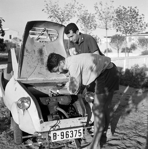 Two young men bend over the open front of a small pale car, its engine exposed beneath the lifted bonnet. Opens a larger view