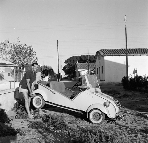 Black-and-white photograph of a man raising the side of a small white open car on rough ground, low white houses behind. Opens a larger view