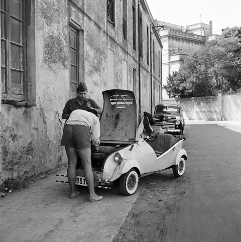 Black-and-white photograph of a small white open car with its bonnet raised by a weathered wall, two men bent over the engine. Opens a larger view