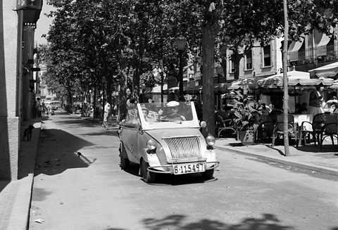 Black-and-white street scene: a small open car with a vertical-bar grille and two occupants comes towards the camera past café tables under plane trees. Opens a larger view