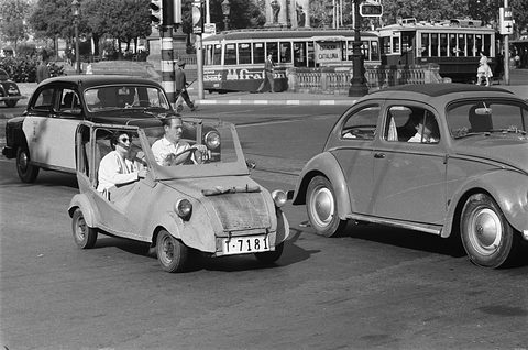Black-and-white photograph of two men in a small open car beside a Volkswagen Beetle and a SEAT saloon, trams behind.