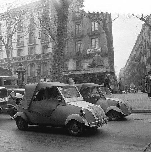 Black-and-white street scene: two small hooded cars with curved vertical-bar grilles parked by a tall building with balconies. Opens a larger view