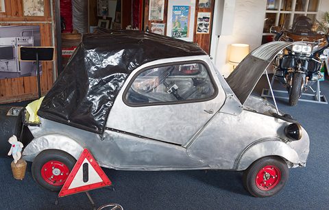 Weathered silver small car with a black hood and red wheels, bonnet open, a warning triangle propped against the front wheel. Opens a larger view