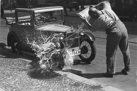 A man in shirtsleeves empties a bucket of water over the bonnet of a small car with its roof folded back parked at a cobbled kerb, spray flying.