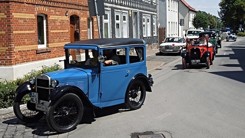 A blue saloon with black wings and wire wheels in a village street, a red tourer and other old cars behind. Opens a larger view