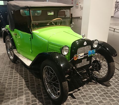 A bright green open tourer with a black hood, black wings and wire wheels on a cobbled museum floor. Opens a larger view