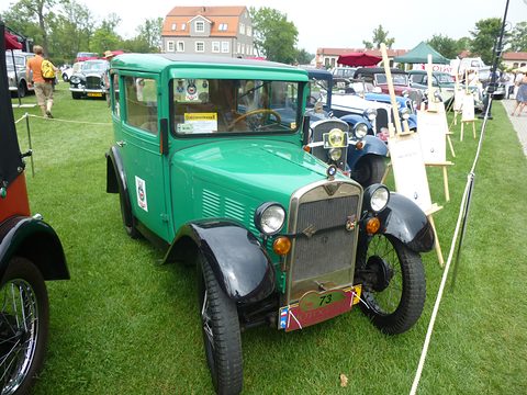 A green saloon with black wings and a chrome radiator parked on grass behind a rope among other old cars. Opens a larger view