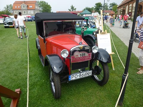 A red open tourer with a black hood and a Motoclassic rally plate parked on grass beside a lamp post. Opens a larger view