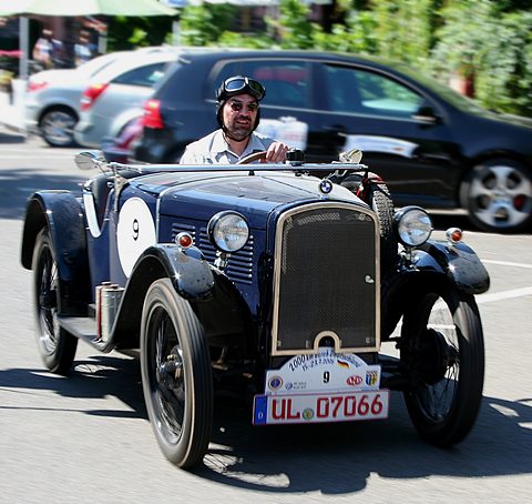 A small dark blue open tourer with a chrome radiator and rally plate, a driver in a leather helmet at the wheel. Opens a larger view