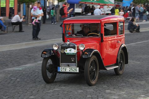A red saloon with black wings and a rally plate on its radiator driving across a cobbled square. Opens a larger view