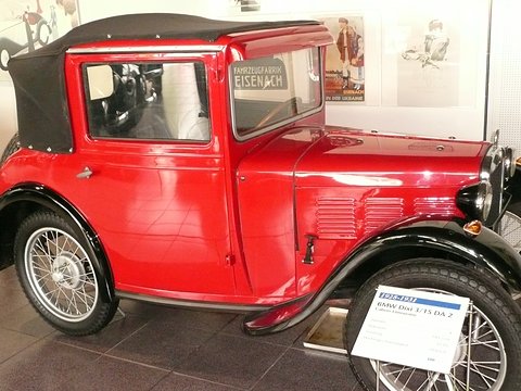 A red two-door car with a black fabric roof, wire wheels and louvred bonnet beside a museum label. Opens a larger view