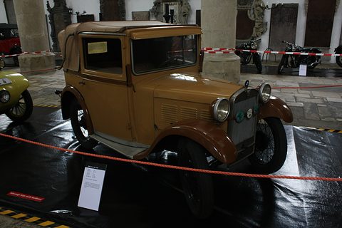 A tan and brown small saloon-cabriolet with a folding roof, behind a red rope on a dark glossy floor. Opens a larger view