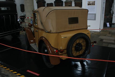 Rear three-quarter view of a tan car with a beige folding roof and a spare wheel on its back. Opens a larger view