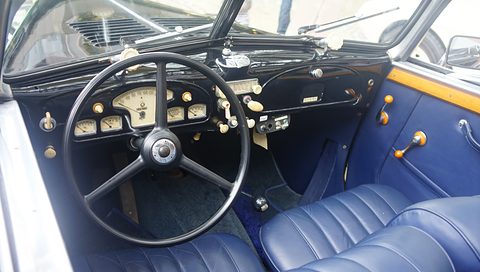 A black four-spoke steering wheel before a cream instrument panel with small gauges, blue pleated seats below. Opens a larger view