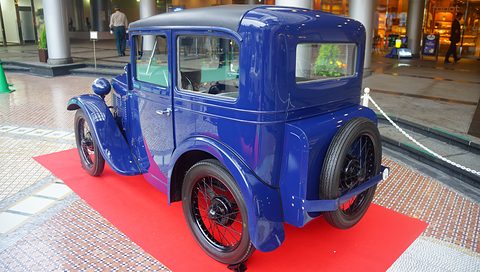Rear three-quarter view of a blue saloon with a spare wire wheel on its tail and red-painted wheel centres. Opens a larger view