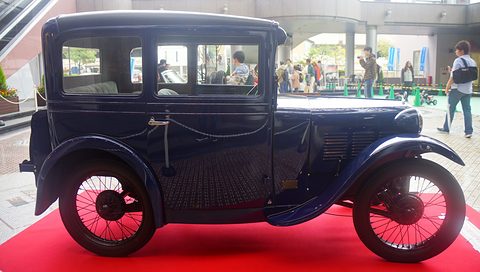 Side view of a tall dark blue two-door saloon with black wings and wire wheels on a red carpet. Opens a larger view