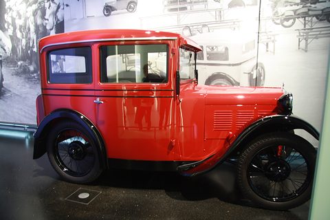 Side view of a red two-door saloon with black wings in front of a large black-and-white photograph mural. Opens a larger view