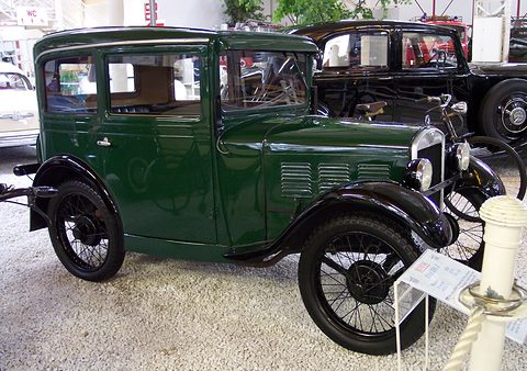 A dark green four-door saloon with black wings and wire wheels on white gravel beside a rope post. Opens a larger view