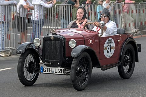 A maroon two-seater with rally numbers driven past barriers by a crew in leather helmets and goggles. Opens a larger view