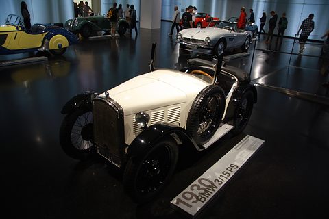 A white two-seater with black wings on a dark floor beside a sign reading 1930 BMW 3/15 PS.