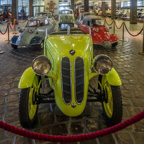Front view of a bright yellow roadster with a split chrome grille and two large lamps on a cobbled floor. Opens a larger view