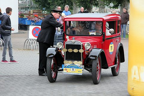 Front view of a red saloon with medallions on its grille and rally plate 11, a uniformed man beside it. Opens a larger view
