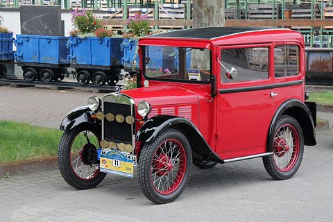 A red two-door saloon with black wings, red wire wheels and rally plaques on its radiator, mine wagons behind. Opens a larger view