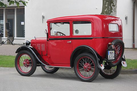Rear three-quarter view of a red saloon with a spare wire wheel and plate on its back, before a white building. Opens a larger view