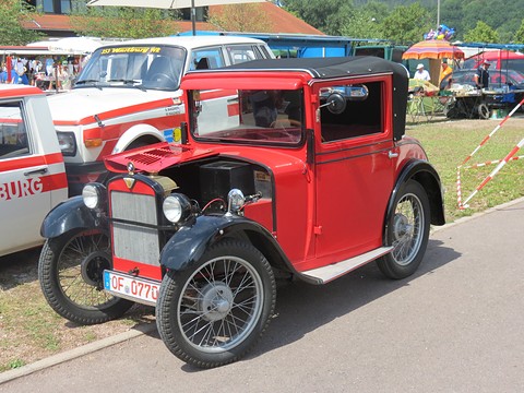 A red saloon with a black roof and wire wheels, its bonnet side raised, beside a red and white car. Opens a larger view