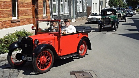 A red open two-seater with black wings and red wire wheels driven along a village street, other old cars behind. Opens a larger view