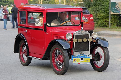 A red two-door saloon with black wings, red wire wheels and medallions on its grille, a driver aboard. Opens a larger view