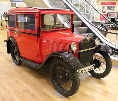 A red two-door saloon with black wings and a BMW DIXI number plate on a wooden floor by an escalator. Opens a larger view