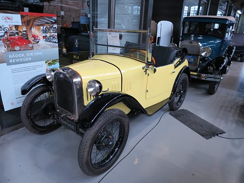 A pale yellow open two-seater with black wings and wire wheels beside a display board, a blue car behind. Opens a larger view