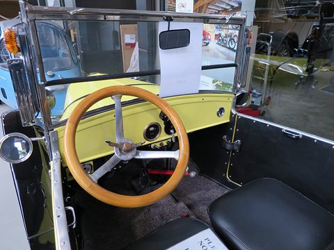 A wooden-rimmed steering wheel before a yellow dashboard with a few gauges, a black seat and a folding windscreen. Opens a larger view