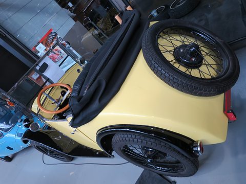 High rear view of a yellow open car with its black hood folded and a spare wire wheel on the tail. Opens a larger view