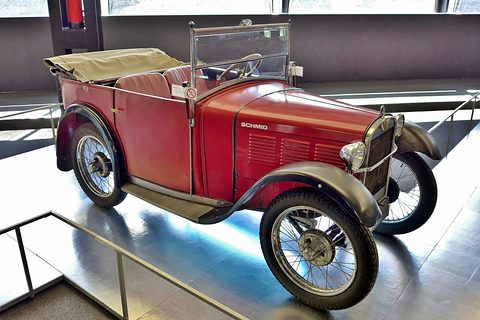 Side view of a red open tourer with a folded beige hood, black wings and wire wheels on a glossy stage. Opens a larger view