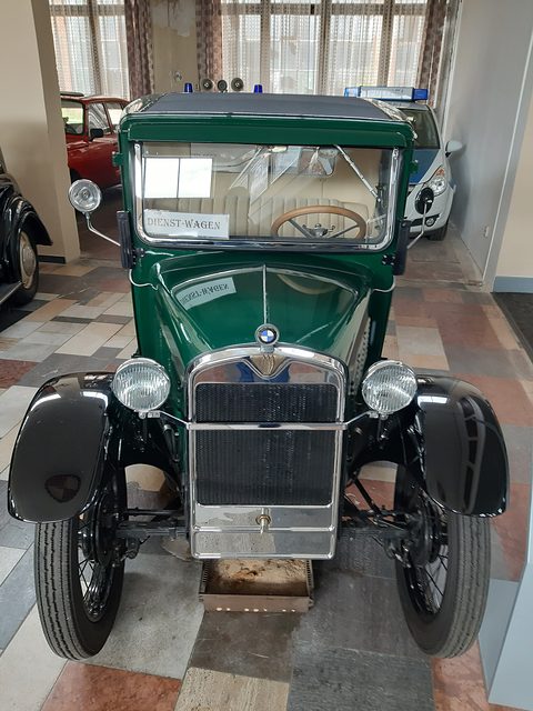Front view of a dark green saloon with a chrome radiator and two headlamps on a tiled museum floor. Opens a larger view
