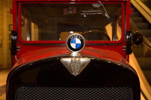 A blue and white roundel on a stalk above a triangular radiator emblem on a red car with a black mesh grille.