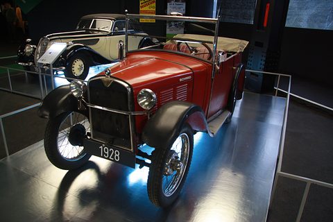 A red open tourer with black wings, a black radiator and a 1928 plate on a lit display stage. Opens a larger view