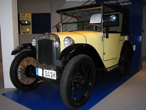 A pale yellow open two-seater with a black hood and plate DA 1H on a blue stand plinth. Opens a larger view