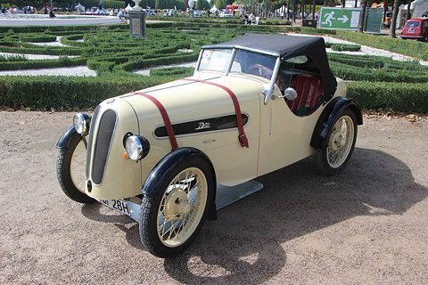A cream two-seater with a black hood, leather bonnet straps and a split grille on gravel by formal hedges. Opens a larger view