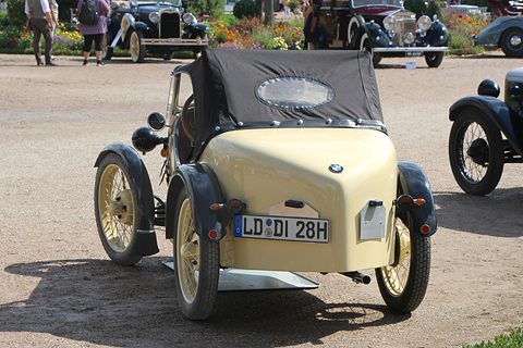 Rear view of a cream two-seater with a rounded tail, a BMW roundel and a black hood on gravel. Opens a larger view