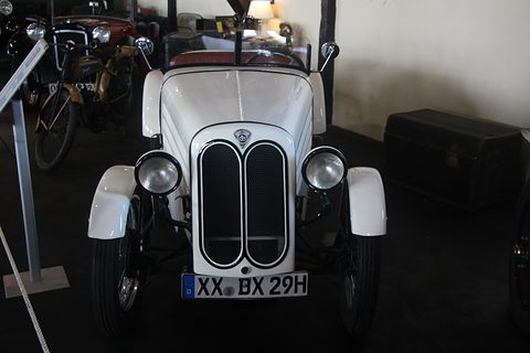 Front view of a white roadster with a black split oval grille and two headlamps in a dim museum hall. Opens a larger view