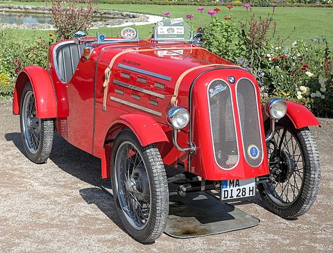 A red two-seat roadster with a split oval grille, leather bonnet straps and wire wheels on gravel before flower beds. Opens a larger view