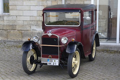 Front view of a maroon saloon with cream wire wheels and black wings on cobbles before a stone wall. Opens a larger view