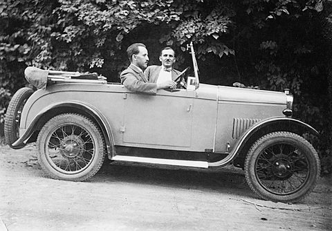 Two men in suits sit in a small open tourer with wire wheels, parked on a gravel road before dense ivy.