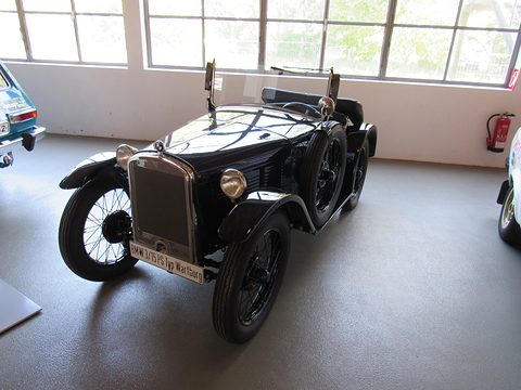 A black open two-seater with a tall radiator and wire wheels on a grey floor by tall windows. Opens a larger view